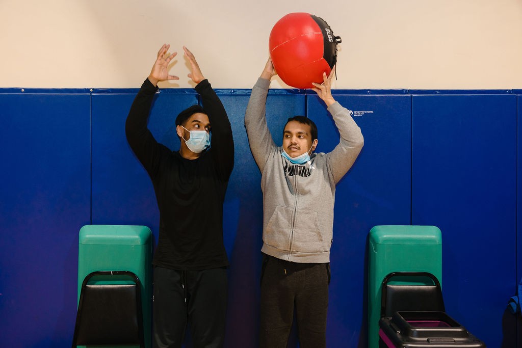 Young male student holding weighted ball over his head as a male fitness instructor shows the student proper lifting technique.