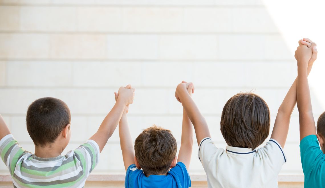 This photo depicts four children from the back, raising their linked hands in a show of unity or celebration. The children are standing against a simple, light-colored background, likely a wall. The two children on the ends are wearing short-sleeved, striped T-shirts, one in green and white and the other in shades of blue. The two children in the middle are wearing solid-colored T-shirts, one in blue and the other in a light color. Their raised hands create a chain, symbolizing friendship, teamwork, or victory. The kids' haircuts and casual clothing suggest an informal, youthful setting, possibly during a playful or educational activity.