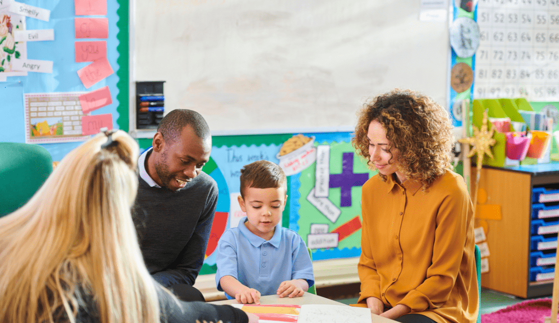 A diverse group of adults and a young child are engaged in an educational activity together in a brightly decorated classroom. The adults are smiling and interacting with the child, creating a warm and supportive environment.