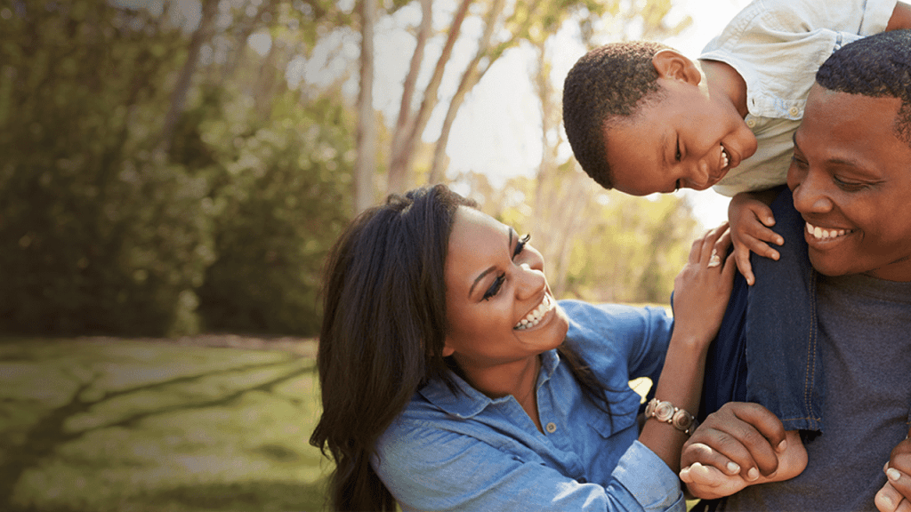 A happy family with a mother, father, and young child enjoying time together outdoors in a sunny park. The child is sitting on the father's shoulders, while the mother looks on with a smile. The background shows green trees and grass.