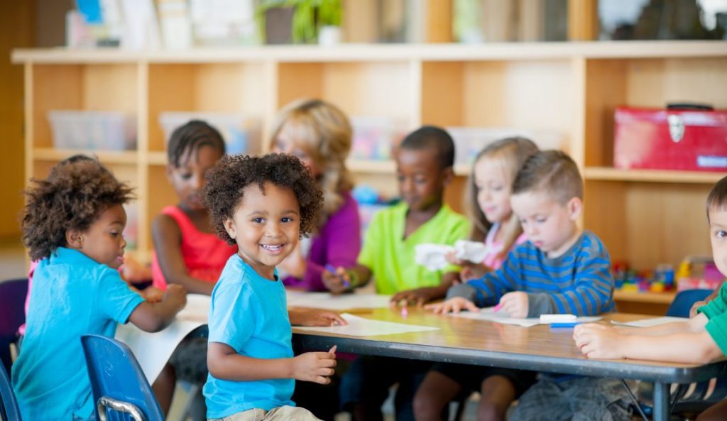 A diverse group of young children sitting around a table in a classroom, engaging in various activities such as drawing and coloring. The children appear happy and engaged, with one child in the foreground smiling brightly at the camera. The background features shelves with educational materials.