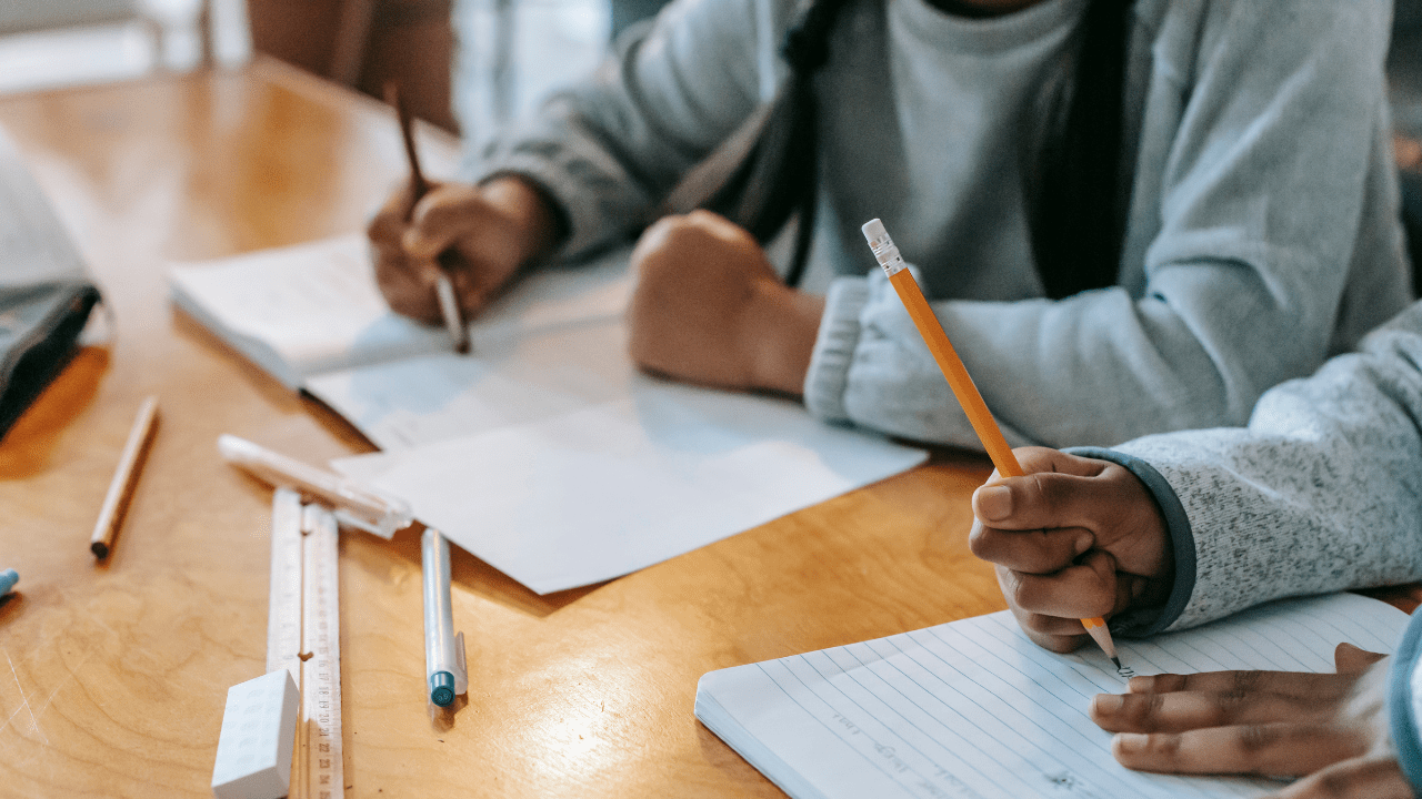 Children writing in notebooks at a wooden table, using pencils and surrounded by rulers and pens.