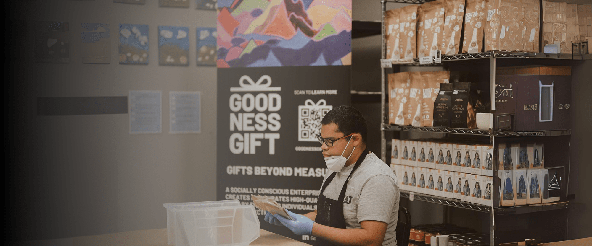 A worker wearing a face mask, gloves, and an apron is carefully packaging products at a table in front of a display that reads 'Goodness Gift: Gifts Beyond Measure.' The display promotes a socially conscious enterprise creating high-quality gifts by individuals on the autism spectrum. Shelves stocked with neatly organized products are visible in the background.