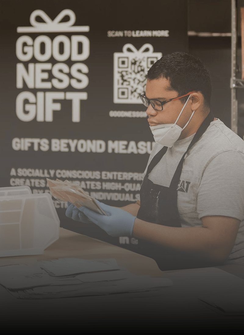 A worker wearing a face mask, gloves, and an apron is carefully packaging products at a table in front of a display that reads 'Goodness Gift: Gifts Beyond Measure.' The display promotes a socially conscious enterprise creating high-quality gifts by individuals on the autism spectrum. Shelves stocked with neatly organized products are visible in the background.