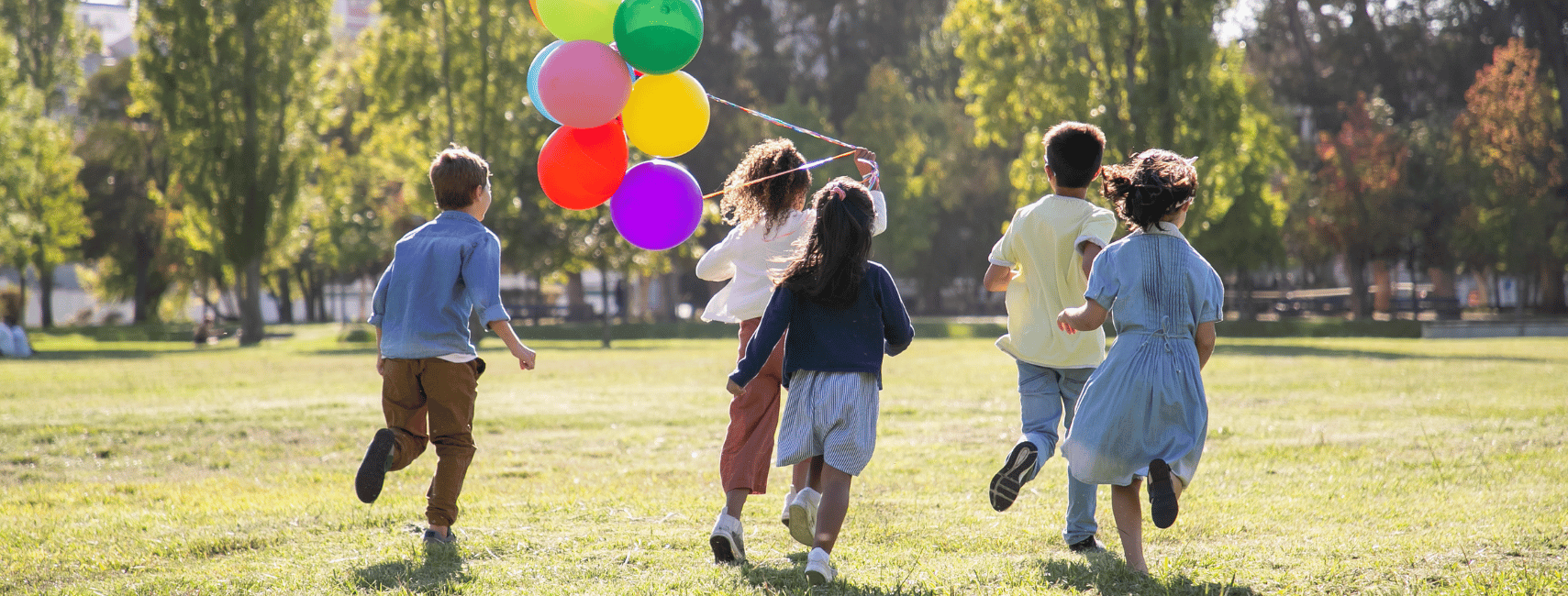 A group of children running together across a grassy park, holding colorful balloons. The scene is bright and joyful, with the children playing under a clear sky, surrounded by green trees, symbolizing unity, happiness, and a sense of community.