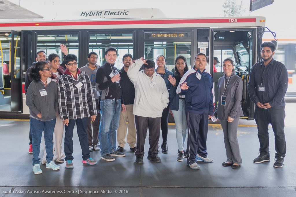 Group of SAAAC Autism Centre students and volunteers/staff standing in front of a TTC hybrid electric bus with a 'To Morningside Hts' sign. The group is smiling and waving, showcasing the supportive and inclusive environment provided by the SAAAC Autism Centre during an accessible transit learning experience in Toronto.