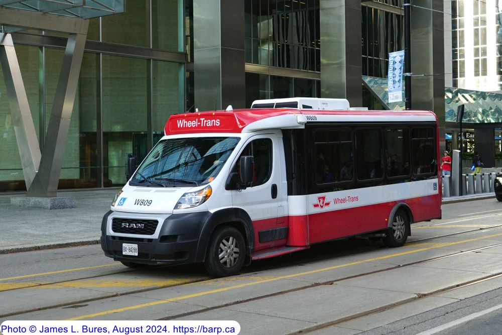 Image of a Toronto Transit Commission (TTC) Wheel-Trans vehicle, a white and red accessible shuttle bus labeled 'Wheel-Trans' on its roof, with vehicle number W809. The bus is parked on a Toronto city street in front of a modern glass building. Wheel-Trans offers accessible transportation for individuals with disabilities, including autism, helping families access autism support services in Toronto.