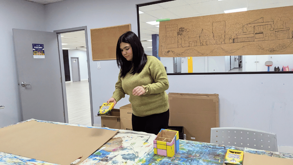 Person preparing an art workspace with craft supplies, including a box of crayons, on a colorful and paint-splattered table. A large blank paper is laid out, with a sketched cityscape pinned on the wall behind them in a creative studio setting.