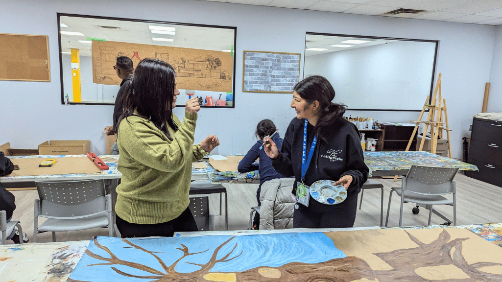 Two individuals engaging in a cheerful conversation while painting in an art studio. One holds a paint palette and brush, and the other gestures animatedly. A painted canvas featuring a tree and blue background is on the table, with other participants working in the background. The room has a creative atmosphere, with sketches on the wall and art supplies on the tables.