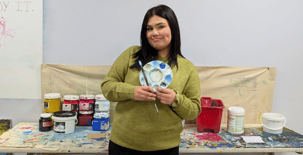 A person holding a paint palette and brush, smiling in front of a colorful art workspace. The background features various paint containers and a canvas, creating a creative and vibrant setting.