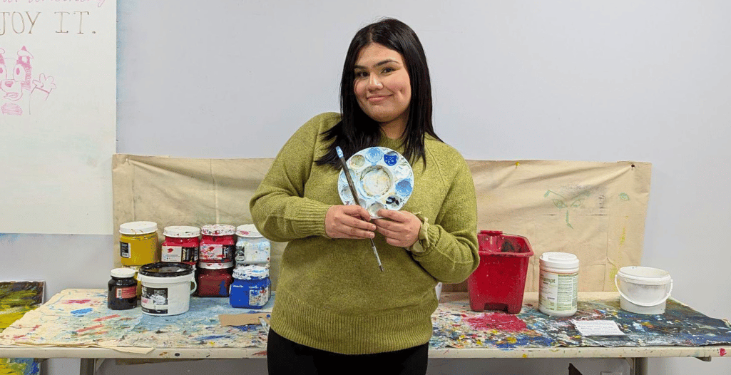 A person holding a paint palette and brush, smiling in front of a colorful art workspace. The background features various paint containers and a canvas, creating a creative and vibrant setting.