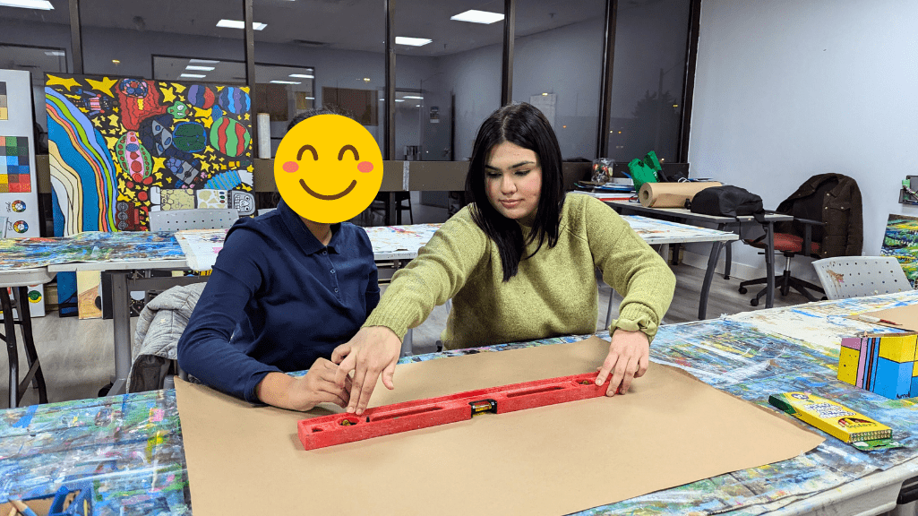 An individual assisting another person in aligning a red level tool on a large sheet of paper in an art studio. The workspace is colorful, featuring a variety of vibrant artwork displayed in the background. The setting has a creative and collaborative atmosphere, with art supplies like crayons visible on the table.