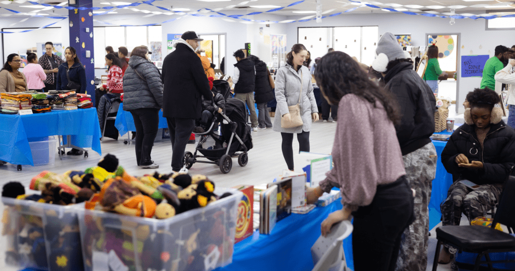 A vibrant scene from SAAAC's Holiday Market, featuring people browsing various stalls with colorful merchandise, including handmade dolls and books, while engaging in a warm and festive atmosphere. Blue decorations adorn the space, and community members of all ages are enjoying the event.