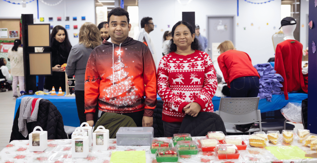 A caregiver and an autistic learner standing behind a table at SAAAC's Holiday Market, proudly showcasing their small business. The table is filled with an assortment of colorful cakes and packaged treats, with festive holiday attire adding to the cheerful atmosphere.