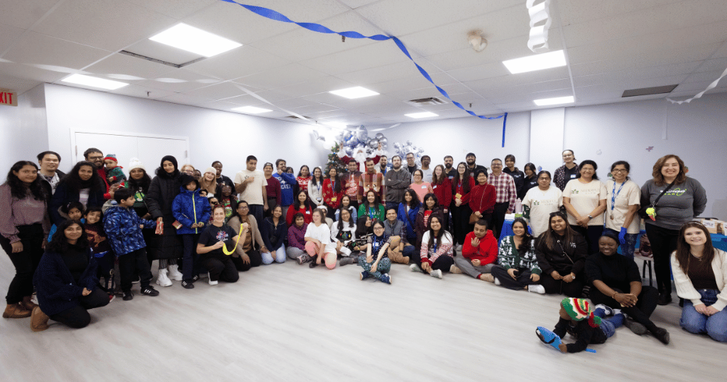 A large group photo taken at SAAAC's Holiday Market, featuring participants, staff, volunteers, and families standing and sitting together in a festive room decorated with blue and white streamers. The group smiles warmly, creating a sense of community and celebration.