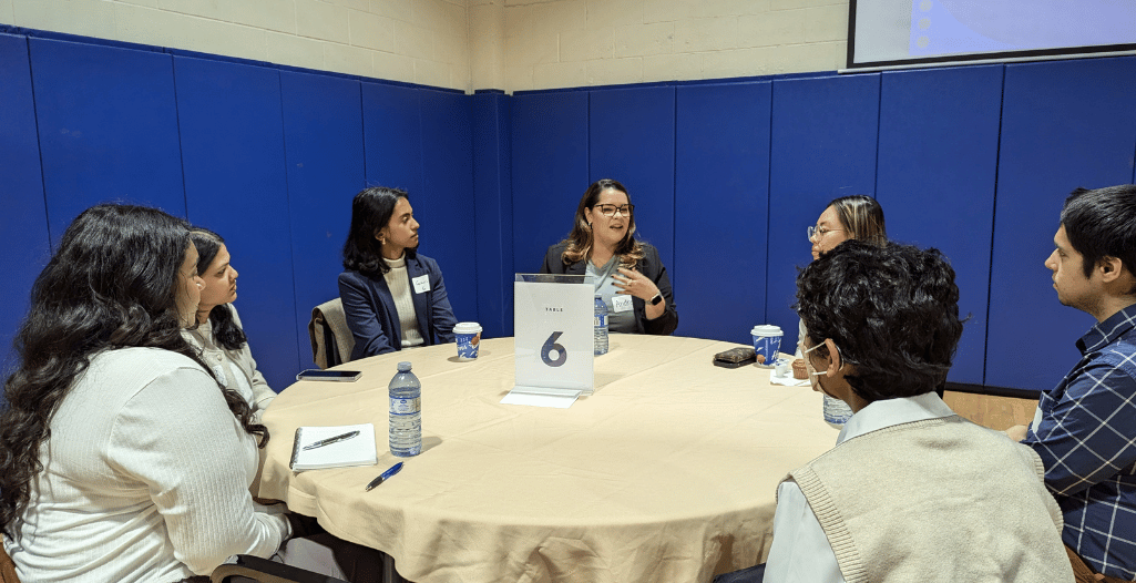 A group of seven individuals seated at a round table with a beige tablecloth during a networking event. A woman in the center speaks animatedly while others listen attentively, with a mix of thoughtful and engaged expressions. A table sign labeled '6' is prominently displayed along with water bottles, notebooks, and Tim Hortons coffee cups. The background features blue padded walls in a gymnasium-like setting, with part of a projected screen visible in the upper corner.