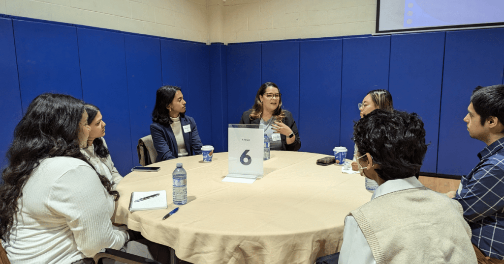 A round table discussion at a networking event, with seven individuals seated around a table covered with a beige tablecloth. A woman in the center is speaking, engaging the group. The table has a card labeled 'Table 6,' water bottles, notebooks, pens, and coffee cups. All participants appear attentive, with some taking notes. The background features blue padded walls in a gymnasium, and a screen is partially visible on the wall.