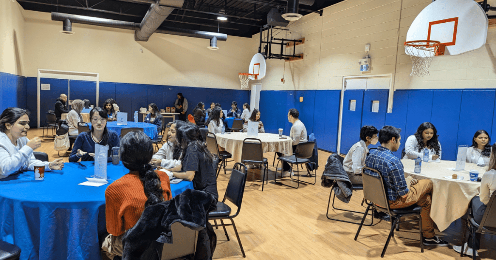 A networking event in a gymnasium, featuring several groups of people seated at round tables covered with blue or beige tablecloths. Attendees are engaged in conversations, with some holding drinks and notepads. The room has blue padded walls, basketball hoops, and a wooden floor. The atmosphere appears lively and collaborative.