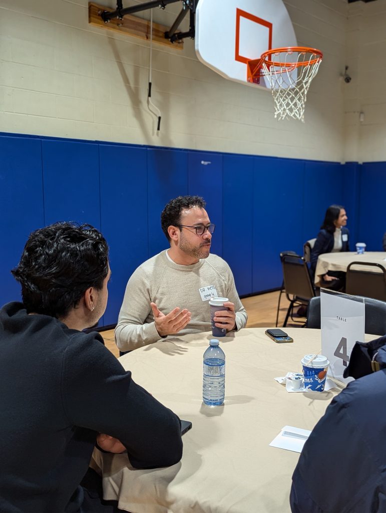 A networking event featuring a round table with a beige tablecloth. At the table, Dr. Jonathan Weiss in a light-colored sweater, wearing glasses and a name tag, gestures as he speaks while holding a coffee cup. Two other individuals are seated, one visible from behind. On the table are a water bottle, another coffee cup, and a number card labeled '4.' In the background, a basketball hoop is mounted on the wall of the gymnasium, along with blue padded walls.