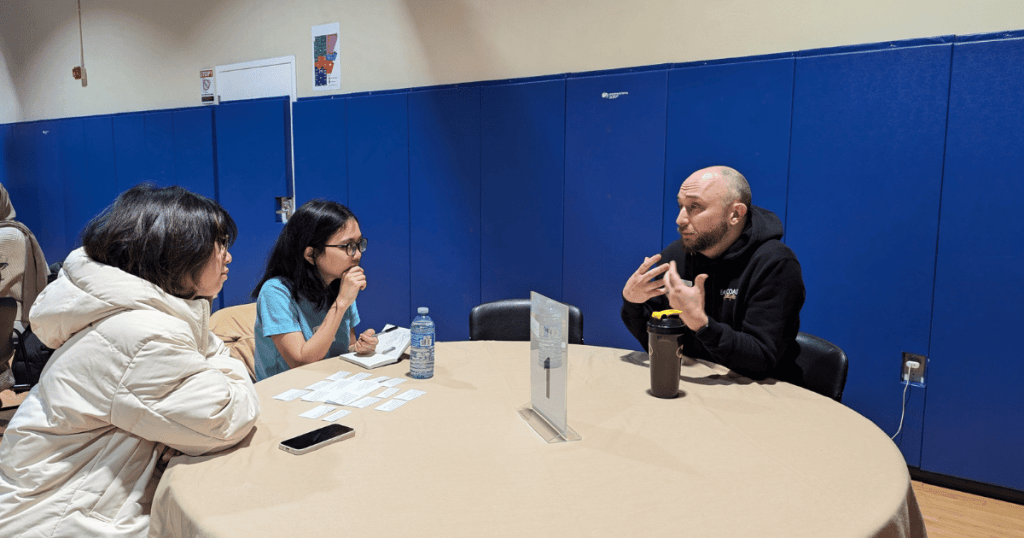 A round table with a beige tablecloth, featuring three individuals engaged in conversation. On the left, two women sit attentively, one in a white winter coat and the other in a light blue shirt, holding a notebook. On the right, a man in a black hoodie gestures expressively as he speaks. The table has a water bottle, a smartphone, and small cards scattered on it. A name card holder stands in the center of the table. The background shows blue padded walls in a gymnasium setting.