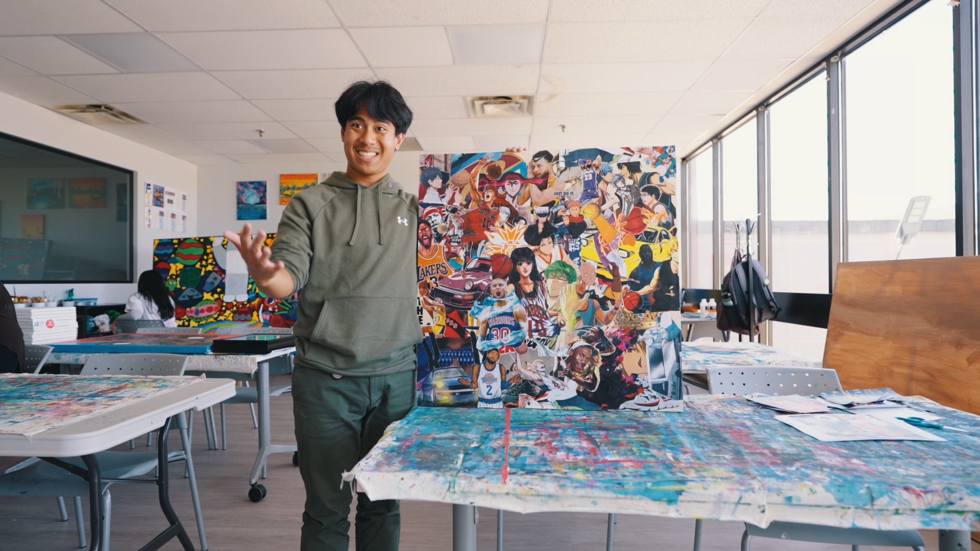 "An artist participating in the SAAAC Autism Centre's Visual Arts Program stands proudly in a creative studio space, holding a vibrant collage artwork featuring various basketball players and pop culture icons. The workspace is filled with colorful art materials and completed artworks, showcasing the program's creativity and inclusivity.