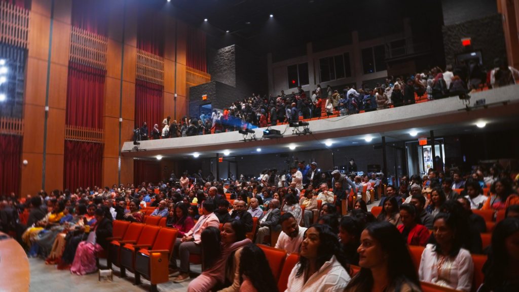 "An auditorium filled with a diverse and vibrant audience attending the Amma, Where Are You? musical hosted by the SAAAC Autism Centre. The photo captures the energy of the event, with attendees seated in bright red chairs, engaging with the performances, and others moving about in the background. The stage and curtains add to the grandeur of the space, showcasing a lively and inclusive gathering