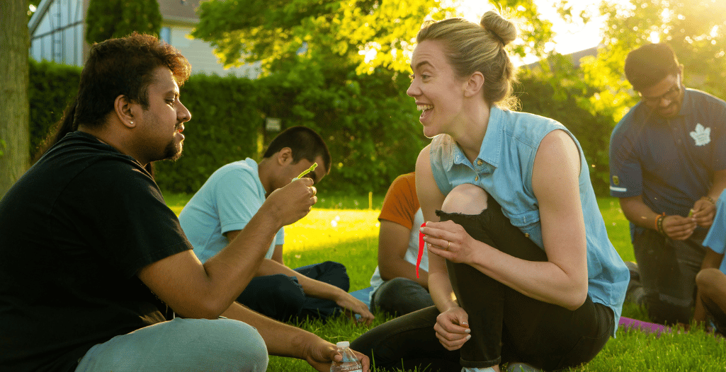 An instructor engaged in a lively outdoor activity with an adult autistic learner, both smiling and interacting in a supportive and inclusive environment, with other participants visible in the background.