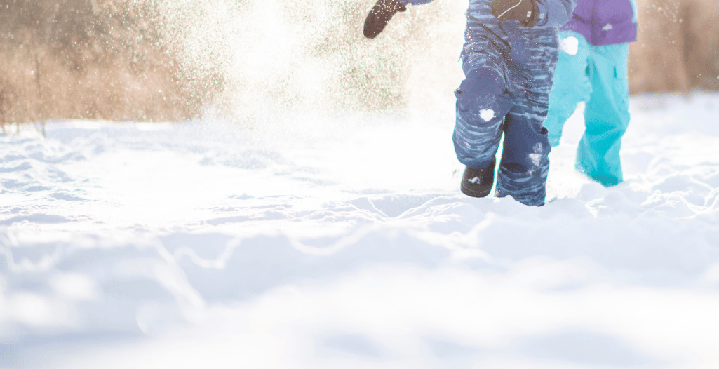 Children playing in the snow on a sunny winter day, dressed in colorful winter gear, with snow flying around as they run.