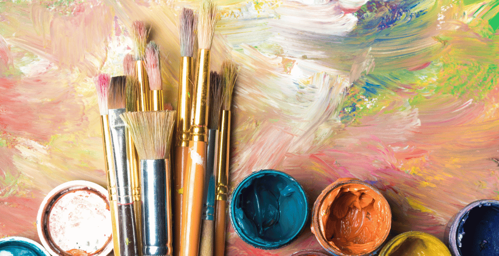 A collection of paintbrushes with colorful paint on their bristles, resting against a vivid, abstractly painted background, accompanied by open paint jars in shades of teal, orange, and white.