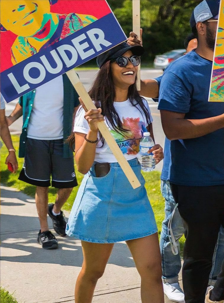 Laksha, a former SAAAC volunteer, participates in the SAAAC Walkathon. She is smiling while holding a colorful protest-style sign that reads 'LOUDER.' She wears a black cap, round sunglasses, a white graphic t-shirt, and a denim skirt. She holds a water bottle in one hand and walks alongside other participants, who are also holding similar signs. The event is outdoors on a sunny day, with a sidewalk and greenery in the background.