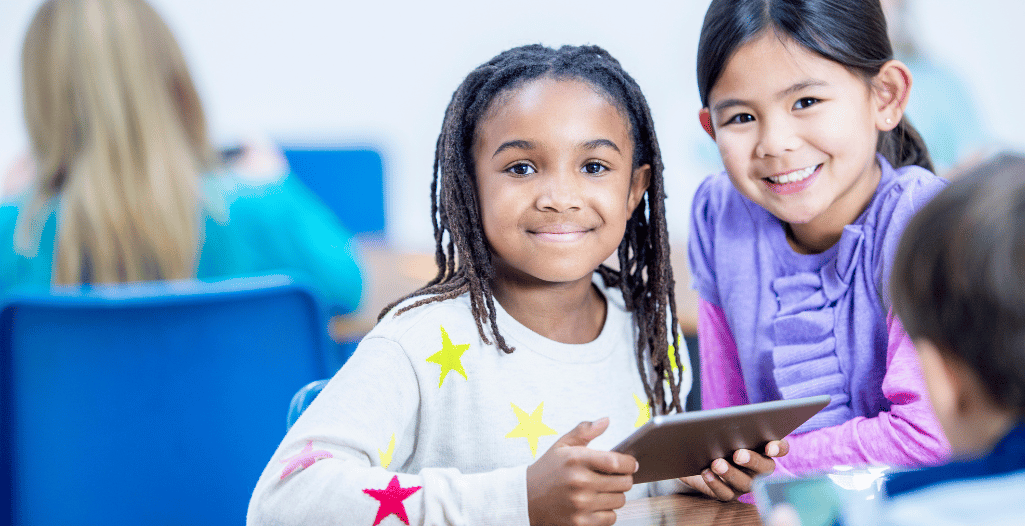 Two young girls smiling and holding a tablet in a classroom setting, with other children in the background.