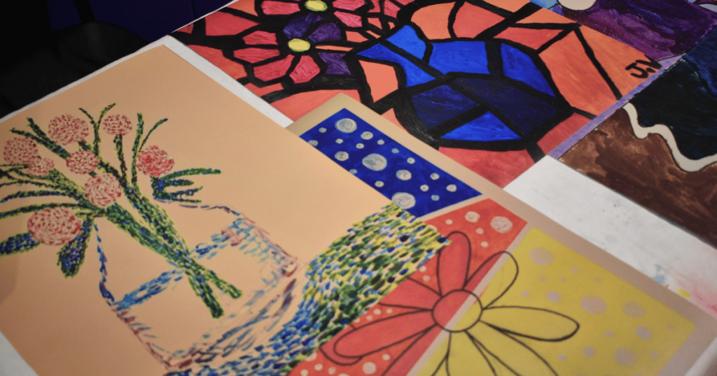 A collection of colourful paintings and drawings created by students in SAAAC’s arts program, laid out side by side on a table.