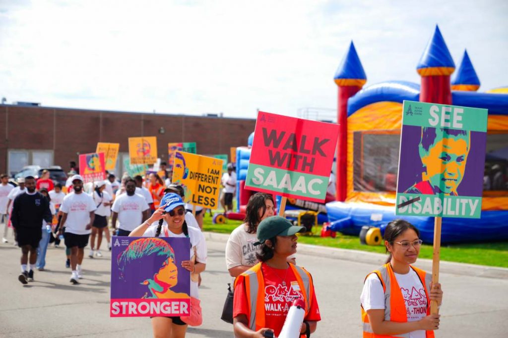 Participants walk together during the SAAAC Walk-a-Thon carrying colourful signs promoting autism acceptance and inclusion. Volunteers in orange safety vests lead the group as families gather near inflatable games during the community celebration.