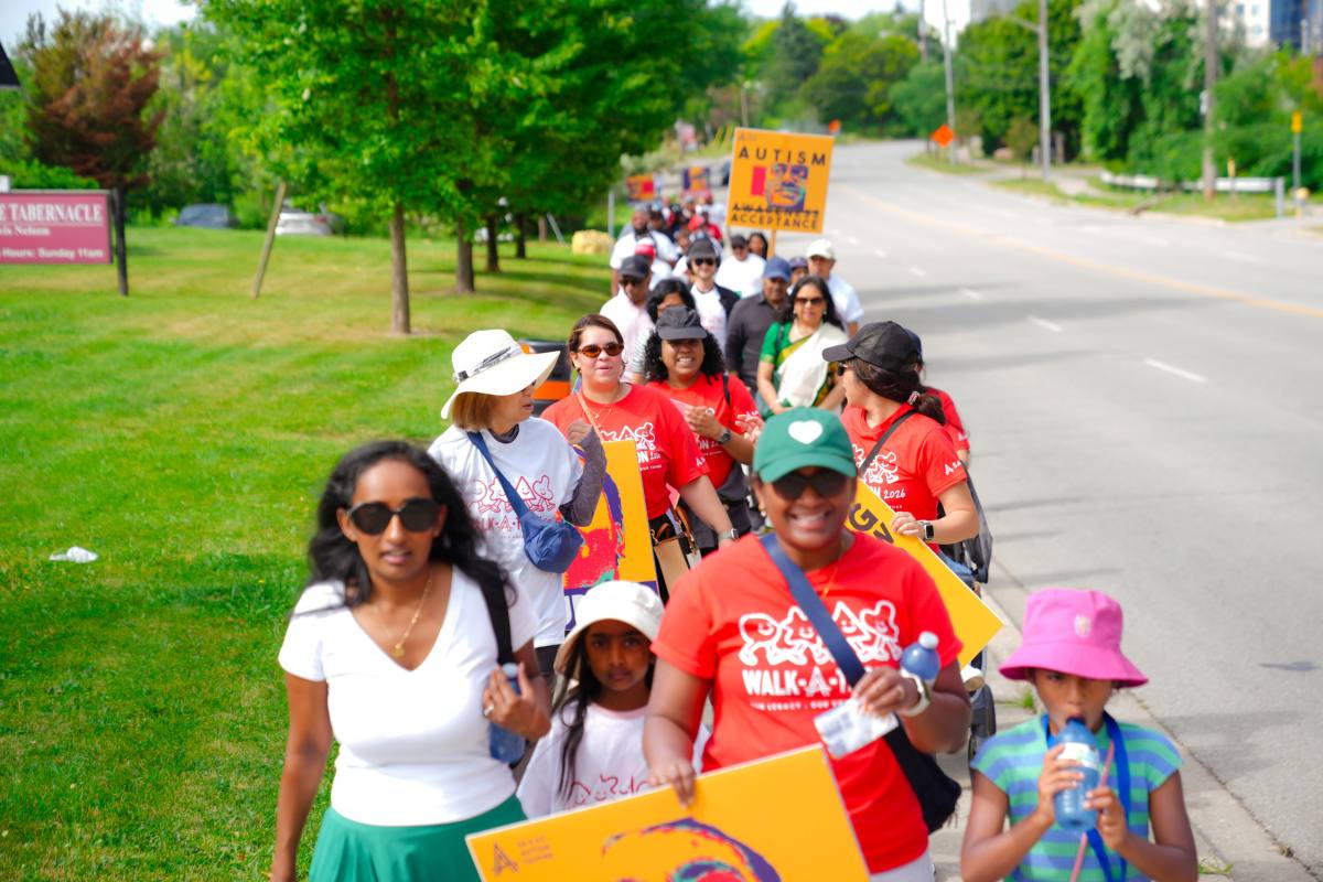 A group of adults and children participate in the SAAAC Autism Centre Walk-A-Thon, walking together along a tree-lined sidewalk on a sunny day. Several participants wear bright red event T-shirts and carry colourful signs promoting autism acceptance and inclusion. The image captures a welcoming, family-friendly community event supporting autism services.